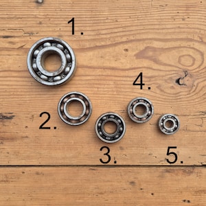 Op de afbeelding: Vijf ronde metalen kogellagers van verschillende groottes op een rij, gelabeld met de cijfers 1, 2, 3, 4 en 5. Elk lager heeft een metalen buitenring, een binnenring en een reeks kleine stalen kogels die in een kooi zijn geplaatst.