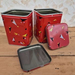 May include: Two square red metal canisters with rounded corners, featuring a scattered pattern of small black, white, and yellow triangles. One canister is open with its matching red lid placed flat in front, while the other lid rests beside it.