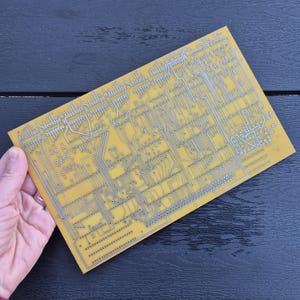 Könnte beinhalten: Eine rechteckige Leiterplatte mit einem komplexen Netzwerk aus silberfarbenen Linien und Komponenten auf gelbem Hintergrund. Die Platine wird von einer Hand gehalten, die ihr kompliziertes Design zeigt. Die Platine wird wahrscheinlich für Elektronik verwendet.