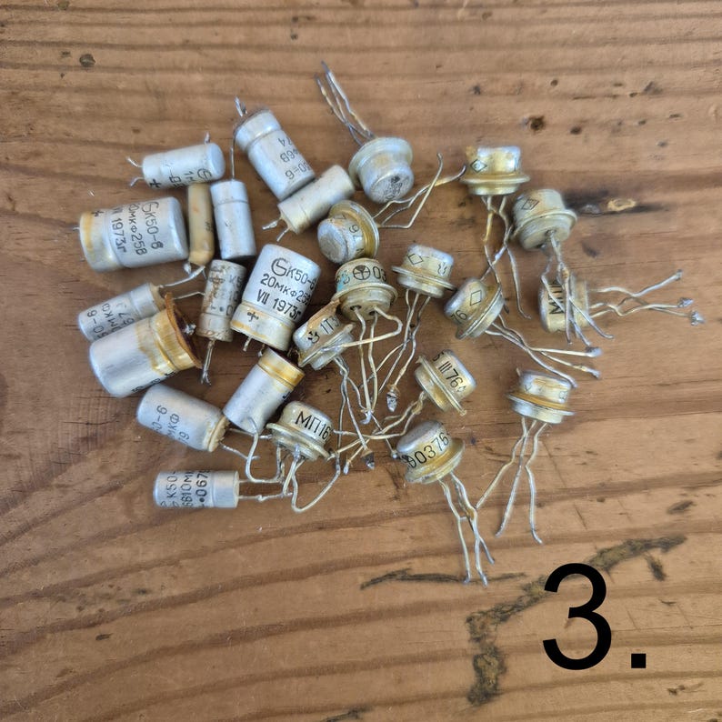 K&ouml;nnte beinhalten: Eine Sammlung alter elektronischer Bauteile, bestehend aus silberfarbenen zylindrischen Kondensatoren und metallischen Transistoren mit Drahtanschl&uuml;ssen. Mehrere Bauteile tragen kyrillische Schriftzeichen und Zahlen. Die Ziffer 3 ist sichtbar.