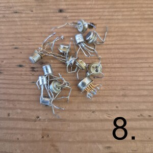 K&ouml;nnte beinhalten: Eine Sammlung kleiner, silberfarbener elektronischer Metalltransistoren mit mehreren d&uuml;nnen Drahtanschl&uuml;ssen, die von ihren Sockeln ausgehen. Einige Bauteile weisen sichtbare alphanumerische Markierungen auf ihren zylindrischen Oberseiten auf. Die Zahl 8 ist in Schwarz aufgedruckt.