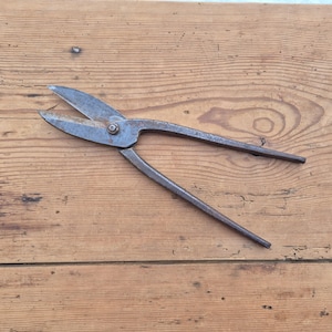 May include: A pair of vintage metal shears, showing signs of age and rust. The blades are open, displaying their sharp edges. The handles are long and slender. The shears are lying on a wooden surface.