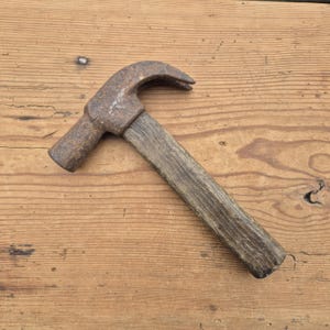 Puede incluir: Un martillo de uña con mango de madera y cabeza de metal oxidado. El martillo está sobre una superficie de madera, mostrando su diseño clásico. Esta herramienta se usa comúnmente en construcción y carpintería.