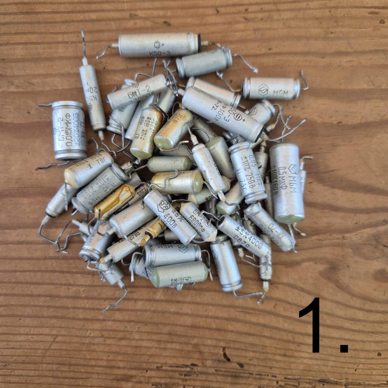 K&ouml;nnte beinhalten: Eine Sammlung verschiedener zylindrischer elektronischer Kondensatoren in Silber-, Grau- und hellen Beiget&ouml;nen. Jedes Bauteil verf&uuml;gt &uuml;ber metallische Drahtanschl&uuml;sse an den Enden sowie aufgedruckte technische Markierungen, einschlie&szlig;lich Text und Symbolen.