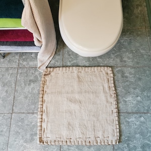 May include: A square, light beige fabric mat featuring a visible whipstitch border in a darker tan thread. The mat is positioned on a gray tiled floor in front of a white toilet, with a metal rack holding folded towels nearby.