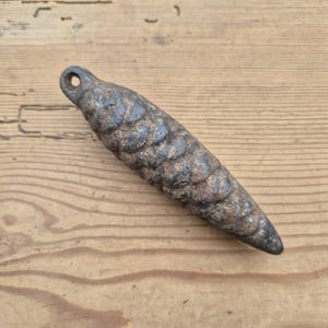 May include: A weathered, cone-shaped metal object with a loop at the top. The surface has a textured, scale-like pattern and a dark, aged appearance. The object is likely a weight or decorative piece.