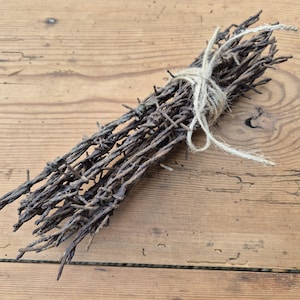 May include: A bundle of weathered, rusty barbed wire tied with twine, resting on a wooden surface. The barbed wire is a dark brown color, with sharp barbs. The twine is a light beige color.