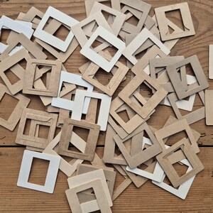 May include: A collection of square cardboard slide mounts in shades of white and light brown. Each mount features a central rectangular cutout designed to hold photographic film. The mounts are scattered in a random overlapping arrangement.