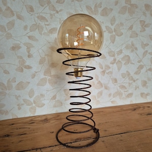 May include: A vintage-style light bulb with a coiled filament sits on a rusty metal spring base. The base is shaped like a traditional bed spring.
