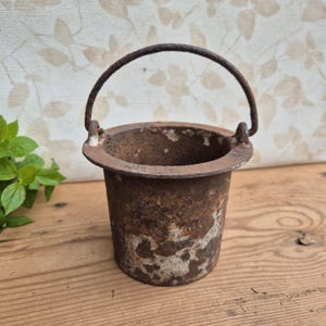 Può includere: Un piccolo secchio cilindrico in ghisa caratterizzato da una patina invecchiata color ruggine e grigio. Il recipiente presenta un bordo largo e piatto e un sottile manico in metallo arcuato fissato a due perni laterali, con segni di ossidazione.