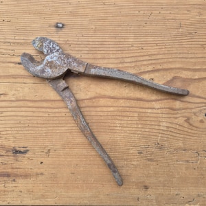 Könnte beinhalten: Eine alte Metall-Zaunzange mit einer verwitterten, rostigen Oberflächenstruktur. Das Werkzeug verfügt über ein gebogenes Backendesign mit einem Drehpunkt und zwei langen, konischen Griffen. Das Metall weist Anzeichen von Oxidation und Abnutzung auf.