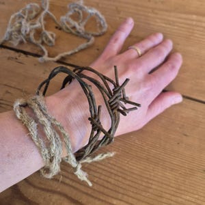 Op de afbeelding: Een rustieke armband gemaakt van verweerd prikkeldraad en vastgebonden met natuurlijk touw. De armband heeft een donkerbruine kleur en is om een pols gewikkeld. Extra touw is zichtbaar op de achtergrond.