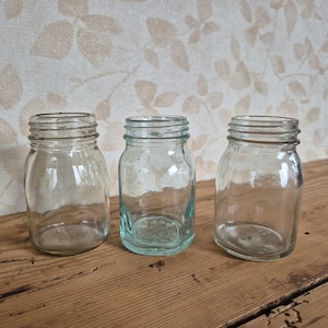 Op de afbeelding: Drie heldere glazen weckpotten met geribbelde schroefdeksels. De potten zijn leeg en staan op een houten oppervlak. De achtergrond heeft een beige bloemenbehang.