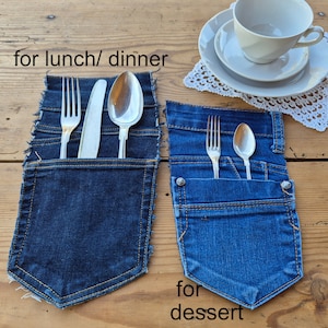 May include: Two denim pocket placemats with silverware. One pocket holds a fork, knife, and spoon, with the text "for lunch/ dinner". The other pocket holds a fork and spoon, with the text "for dessert". A teacup and saucer are in the background.
