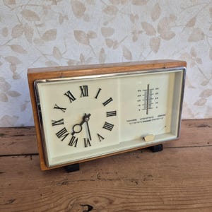 May include: A vintage wooden clock with a cream-colored face, featuring black Roman numerals and hands. The clock also includes a thermometer. The clock is rectangular and sits on two black feet.