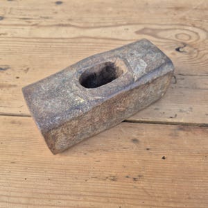 May include: A weathered, rectangular sledgehammer with a large, oval eye. The metal head shows signs of age and use, with a dark, rusty brown color. The tool is designed for heavy-duty tasks.