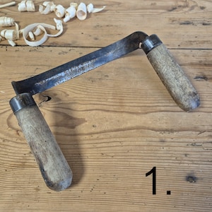 Può includere: Un coltello da trazione vintage con una lama curva e due manici in legno. La lama mostra segni di usura e i manici sono in legno invecchiato. Trucioli di legno sono sparsi intorno all'utensile, suggerendo un uso recente. L'utensile è su una superficie di legno.