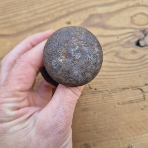May include: A round, dark brown metal knob with a textured surface. The knob is held in a hand against a wooden background. The knob appears to be made of iron or a similar material, and shows signs of age and wear.