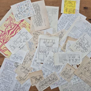 May include: A collection of vintage circuit diagrams and schematics, featuring intricate line drawings in black ink on aged, cream-colored paper. Some diagrams have red and yellow accents. The papers are scattered on a wooden surface.