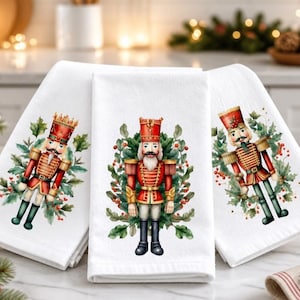 May include: Three white kitchen towels featuring a printed illustration of a nutcracker soldier dressed in a red and gold uniform. Each nutcracker is surrounded by a wreath of green holly leaves and red berries, displayed in a folded arrangement.