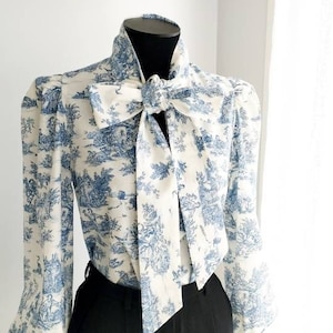 Peut inclure: Un chemisier blanc à motif toile bleu et blanc. Le chemisier a un col haut et un grand nœud au cou. Le chemisier est porté avec un pantalon noir.