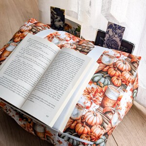 K&ouml;nnte beinhalten: Ein Buchkissen aus Stoff mit einem Muster aus orangefarbenen K&uuml;rbissen, Herbstbl&auml;ttern und Tassen mit Hei&szlig;getr&auml;nken. Ein aufgeschlagenes Buch liegt auf dem Kissen und wird von einem wei&szlig;en Gummiband gehalten. Ein dunkles Tablet steckt in der Tasche.
