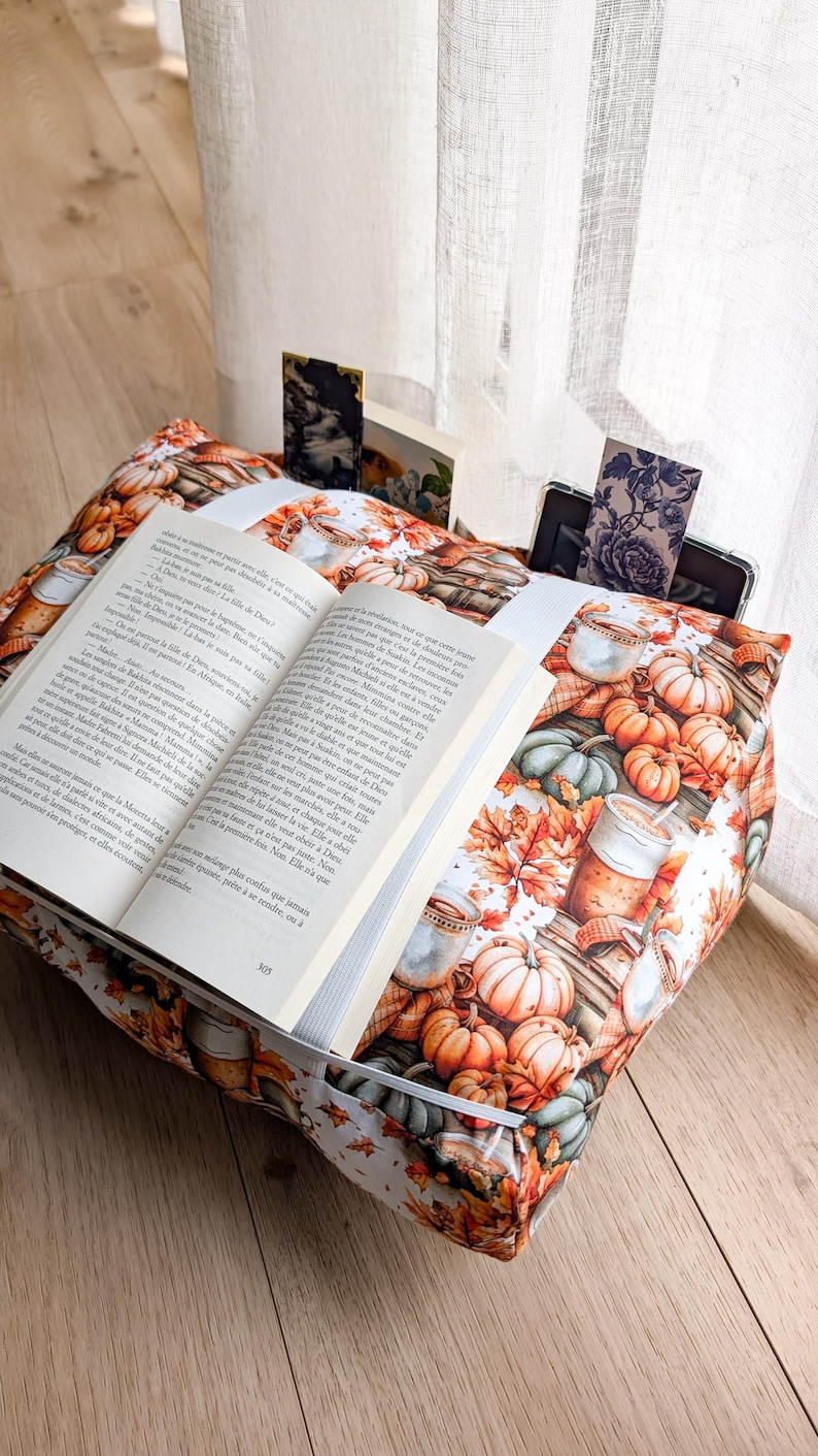 K&ouml;nnte beinhalten: Ein Buchkissen aus Stoff mit einem Muster aus orangefarbenen K&uuml;rbissen, Herbstbl&auml;ttern und Tassen mit Hei&szlig;getr&auml;nken. Ein aufgeschlagenes Buch liegt auf dem Kissen und wird von einem wei&szlig;en Gummiband gehalten. Ein dunkles Tablet steckt in der Tasche.