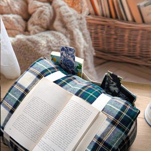 K&ouml;nnte beinhalten: Ein weiches, quadratisches Buchkissen mit einem dunkelgr&uuml;nen, blauen und wei&szlig;en Schottenkaro-Muster. Das Kissen verf&uuml;gt &uuml;ber eine integrierte Tasche f&uuml;r ein Smartphone und einen Schlitz f&uuml;r ein offenes Buch sowie zwei Stoff-Lesezeichen am Rand.