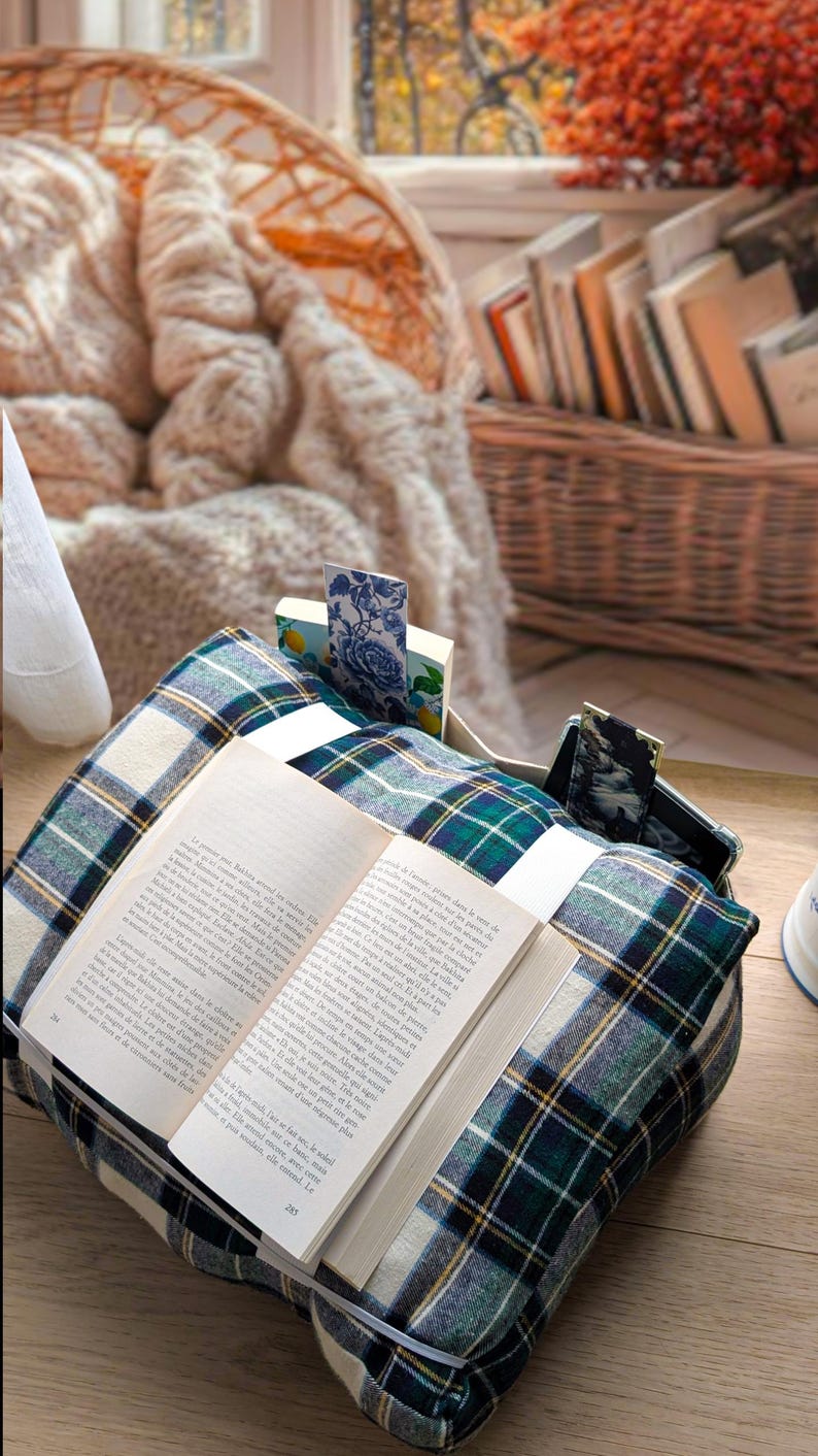 K&ouml;nnte beinhalten: Ein weiches, quadratisches Buchkissen mit einem dunkelgr&uuml;nen, blauen und wei&szlig;en Schottenkaro-Muster. Das Kissen verf&uuml;gt &uuml;ber eine integrierte Tasche f&uuml;r ein Smartphone und einen Schlitz f&uuml;r ein offenes Buch sowie zwei Stoff-Lesezeichen am Rand.