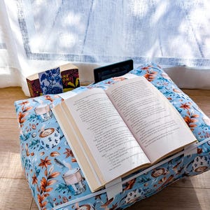 K&ouml;nnte beinhalten: Ein hellblaues Buchkissen aus Stoff mit einem Muster aus Kaffeetassen und Herbstbl&auml;ttern. Das Kissen hat ein wei&szlig;es Gummiband, das ein offenes Buch h&auml;lt, sowie einen kleinen schwarzen E-Reader und ein Buch mit Blumenmuster im Hintergrund.
