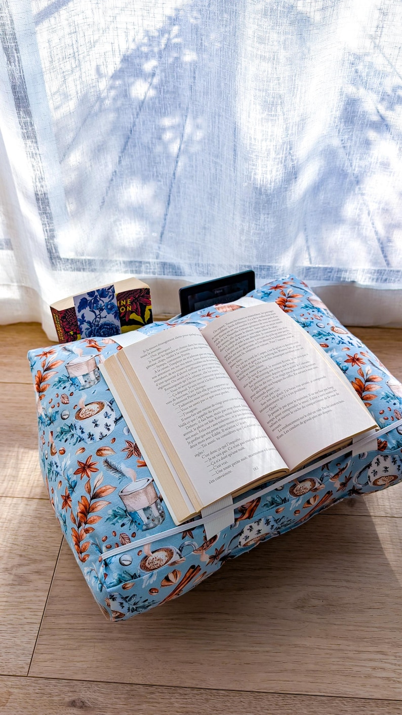 K&ouml;nnte beinhalten: Ein hellblaues Buchkissen aus Stoff mit einem Muster aus Kaffeetassen und Herbstbl&auml;ttern. Das Kissen hat ein wei&szlig;es Gummiband, das ein offenes Buch h&auml;lt, sowie einen kleinen schwarzen E-Reader und ein Buch mit Blumenmuster im Hintergrund.