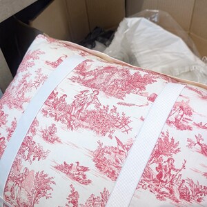 K&ouml;nnte beinhalten: Ein rechteckiges Kissen aus cremefarbenem Stoff mit einem roten Toile-Muster, das pastorale Szenen mit Figuren, B&auml;umen und V&ouml;geln zeigt. Zwei breite, wei&szlig;e elastische B&auml;nder sind horizontal &uuml;ber die Vorderseite des Kissens gespannt.