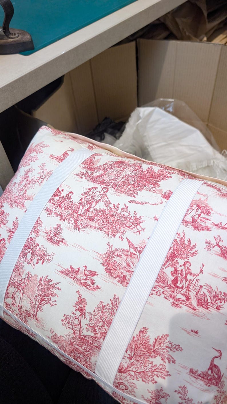 K&ouml;nnte beinhalten: Ein rechteckiges Kissen aus cremefarbenem Stoff mit einem roten Toile-Muster, das pastorale Szenen mit Figuren, B&auml;umen und V&ouml;geln zeigt. Zwei breite, wei&szlig;e elastische B&auml;nder sind horizontal &uuml;ber die Vorderseite des Kissens gespannt.