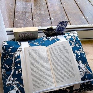 K&ouml;nnte beinhalten: Ein dunkelblauer Buchst&auml;nder aus Stoff mit wei&szlig;em Blumen- und Vogelmuster. Der St&auml;nder h&auml;lt ein aufgeschlagenes Buch mit franz&ouml;sischem Text und verf&uuml;gt &uuml;ber ein wei&szlig;es Gummiband. Dahinter liegt ein Buch mit dem Titel Bakhita von V&eacute;ronique Olmi.