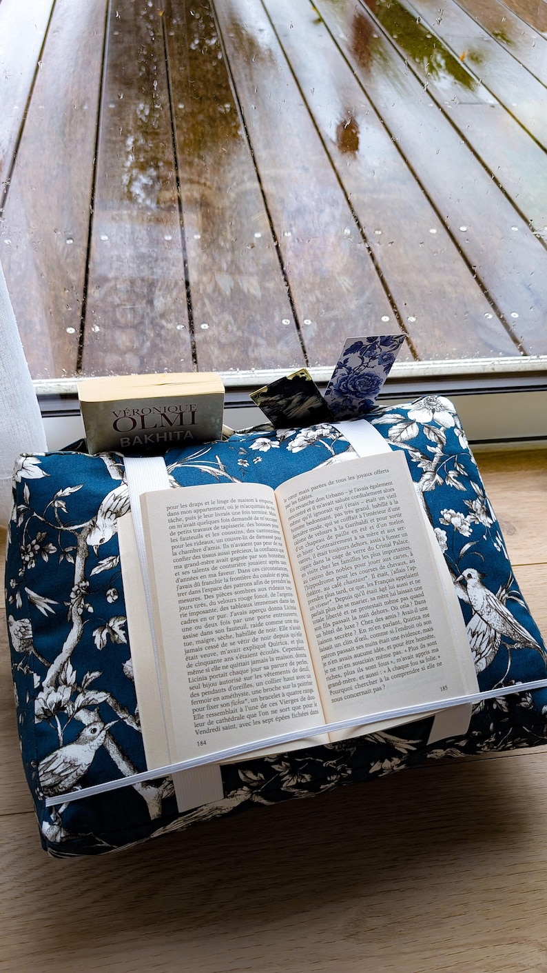 K&ouml;nnte beinhalten: Ein dunkelblauer Buchst&auml;nder aus Stoff mit wei&szlig;em Blumen- und Vogelmuster. Der St&auml;nder h&auml;lt ein aufgeschlagenes Buch mit franz&ouml;sischem Text und verf&uuml;gt &uuml;ber ein wei&szlig;es Gummiband. Dahinter liegt ein Buch mit dem Titel Bakhita von V&eacute;ronique Olmi.