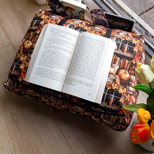 K&ouml;nnte beinhalten: Ein rechteckiges Buchkissen aus Stoff mit einem Aufdruck von B&uuml;cherregalen, K&uuml;rbissen und Herbstbl&auml;ttern. Das Kissen verf&uuml;gt &uuml;ber ein schwarzes Gummiband in der Mitte, das ein offenes Buch h&auml;lt, sowie zus&auml;tzliche Schlitze f&uuml;r Lesezeichen.