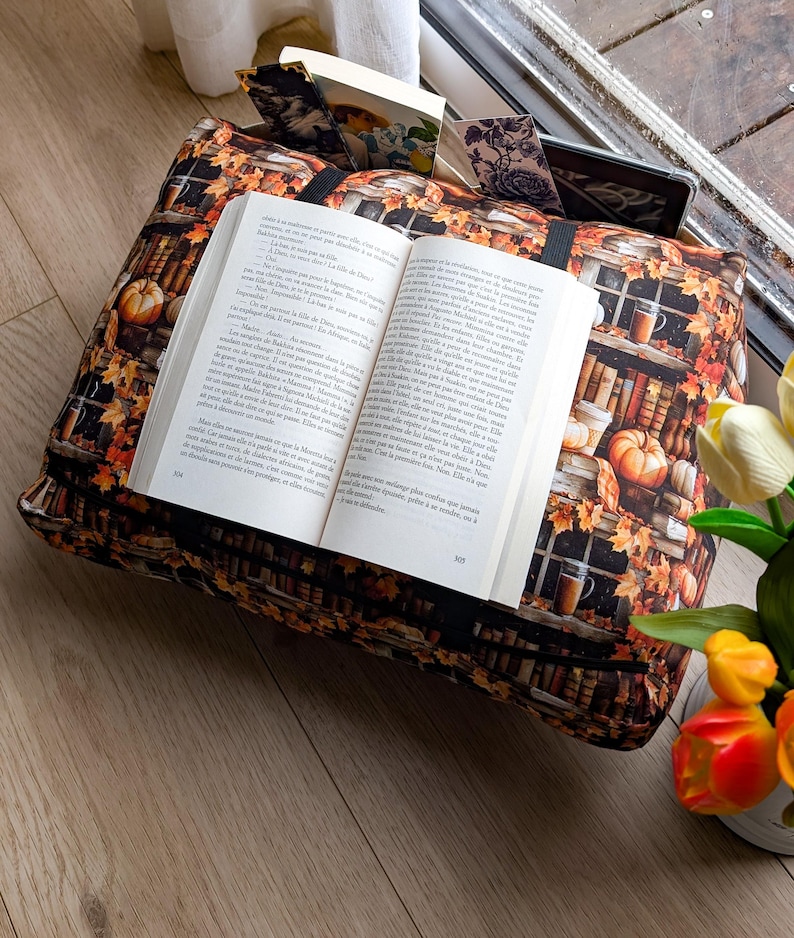 K&ouml;nnte beinhalten: Ein rechteckiges Buchkissen aus Stoff mit einem Aufdruck von B&uuml;cherregalen, K&uuml;rbissen und Herbstbl&auml;ttern. Das Kissen verf&uuml;gt &uuml;ber ein schwarzes Gummiband in der Mitte, das ein offenes Buch h&auml;lt, sowie zus&auml;tzliche Schlitze f&uuml;r Lesezeichen.