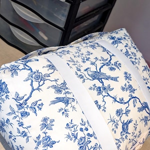 K&ouml;nnte beinhalten: Ein rechteckiges Kissen mit wei&szlig;em Stoffgrund und einem blauen Toile-Muster, das V&ouml;gel auf bl&uuml;henden Zweigen zeigt. Das Kissen wird durch zwei breite, wei&szlig;e Gummib&auml;nder zusammengehalten, die quer um das Kissen gewickelt sind.