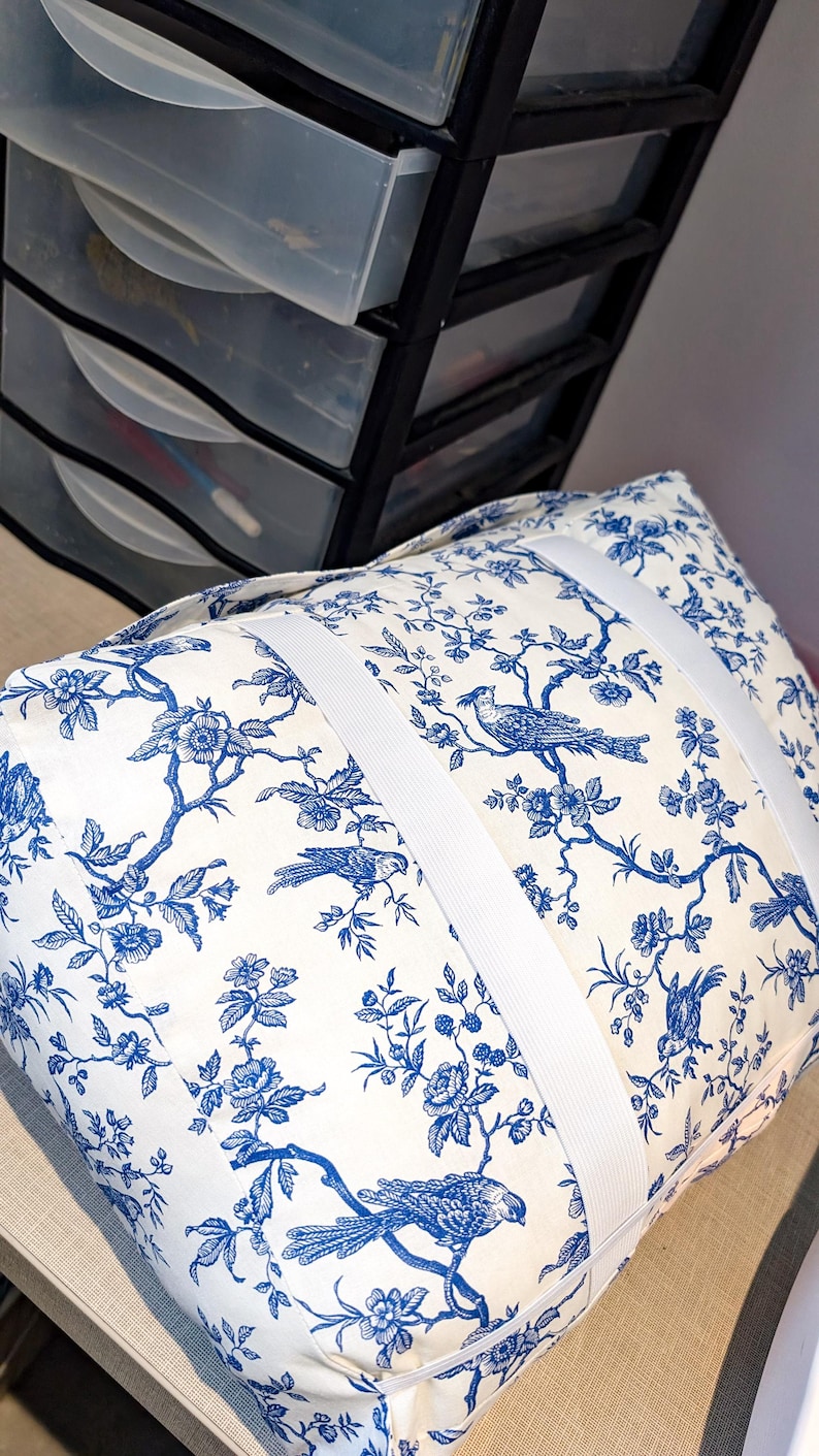 K&ouml;nnte beinhalten: Ein rechteckiges Kissen mit wei&szlig;em Stoffgrund und einem blauen Toile-Muster, das V&ouml;gel auf bl&uuml;henden Zweigen zeigt. Das Kissen wird durch zwei breite, wei&szlig;e Gummib&auml;nder zusammengehalten, die quer um das Kissen gewickelt sind.