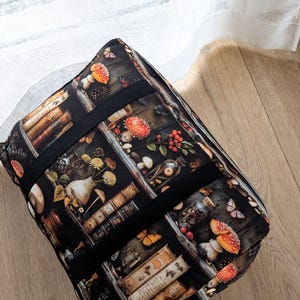 K&ouml;nnte beinhalten: Eine rechteckige Stofftasche mit einem dunklen Aufdruck von B&uuml;cherregalen, rot-wei&szlig;en Pilzen, Glasgef&auml;&szlig;en und Schmetterlingen. Die Tasche hat schwarze Stoffriemen, die &uuml;ber die Oberseite und die Seiten verlaufen, sowie eine strukturierte Form.
