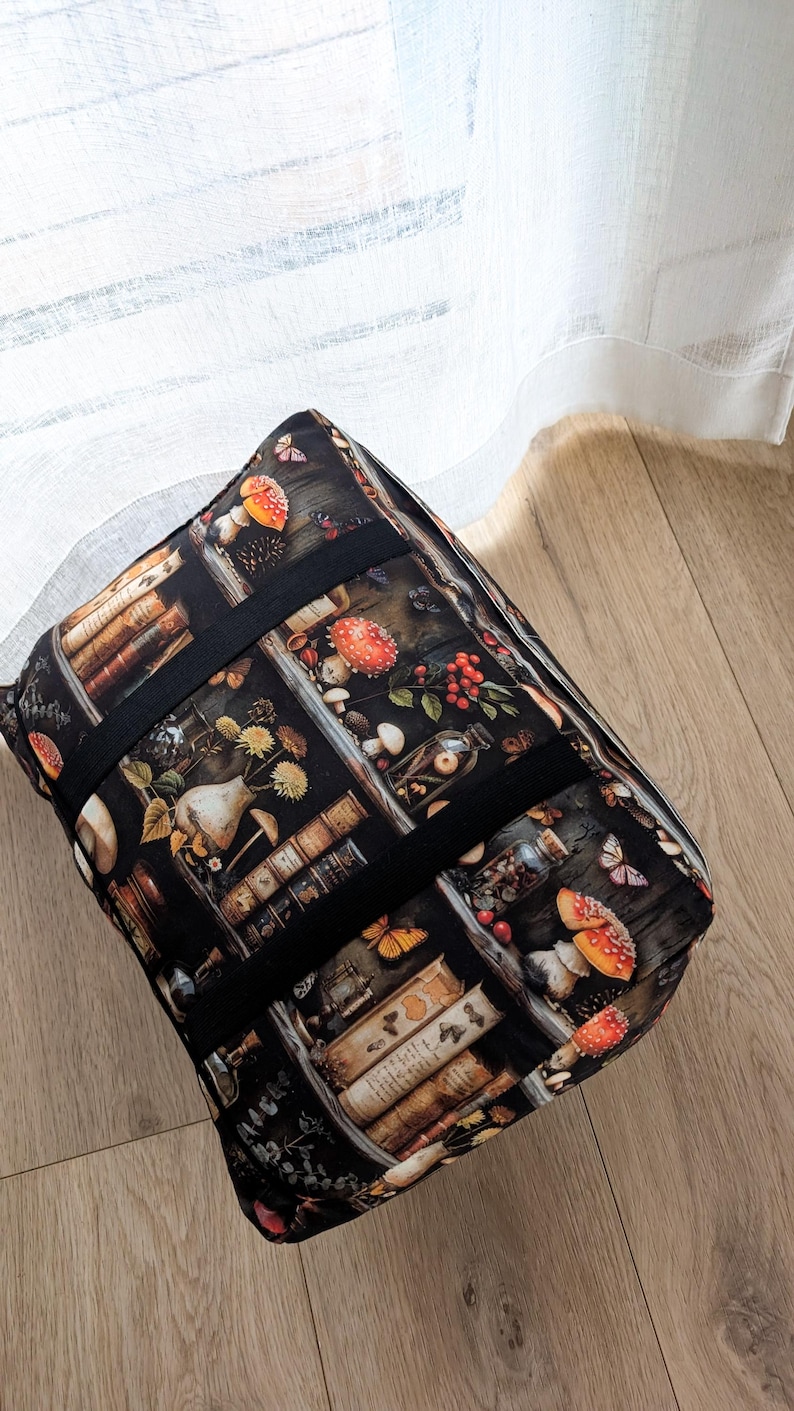 K&ouml;nnte beinhalten: Eine rechteckige Stofftasche mit einem dunklen Aufdruck von B&uuml;cherregalen, rot-wei&szlig;en Pilzen, Glasgef&auml;&szlig;en und Schmetterlingen. Die Tasche hat schwarze Stoffriemen, die &uuml;ber die Oberseite und die Seiten verlaufen, sowie eine strukturierte Form.