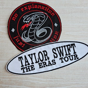 May include: Two embroidered patches. The first is round and black with a red snake with a red tongue and the text "No Explanation Is Required". The second is oval and white with the text "Taylor Swift The Eras Tour".