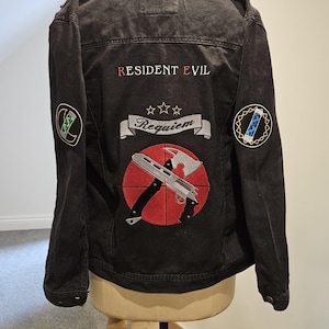 May include: Black denim jacket with embroidered Resident Evil Requiem design. Features a red target with a silver axe and gun, and patches on the sleeves. The jacket is displayed on a mannequin.