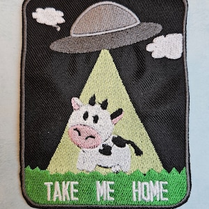 May include: Embroidered patch with a cartoon cow being abducted by a UFO. The design features a black background, a green field, and the text "TAKE ME HOME" in white.