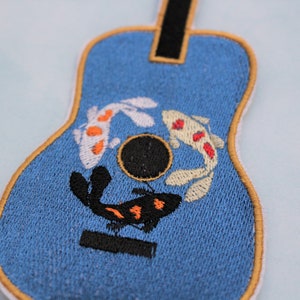 May include: A blue embroidered patch shaped like a guitar with three koi fish swimming around a sound hole. The fish are white with orange and red accents.