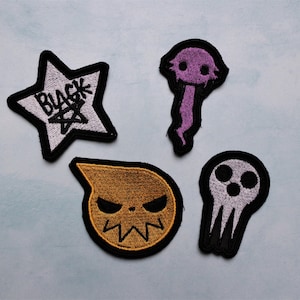 May include: Four embroidered iron-on patches featuring characters from the anime Soul Eater. The patches are white, purple, yellow, and grey. The patches feature the words "Black*" and a star, a ghost-like figure, a yellow character with a frown, and a skull with three eyes.