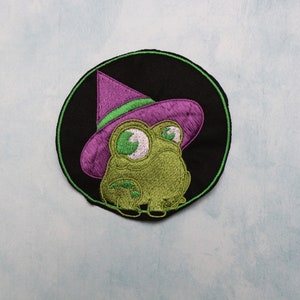 May include: Embroidered patch featuring a green frog wearing a purple witch hat with a green brim. The frog has large, expressive eyes and is set against a black circular background with a green border. The patch is on a light blue surface.