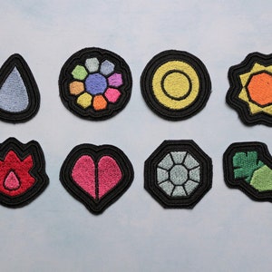 May include: Eight embroidered patches with black edging. The patches display diverse colourful designs, including a teardrop, a flower, a sun, and geometric shapes. The patches are likely for clothing or accessories, and are measured in centimetres.