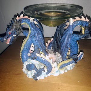 Könnte beinhalten: Ein dekorativer Kerzenhalter mit Fantasy-Thema, der zwei blaue Drachen mit ausgebreiteten Flügeln zeigt, die eine klare Glasschale tragen. Die Drachen haben detaillierte Schuppen und rote Augen und stehen auf einer Wolkenbasis.