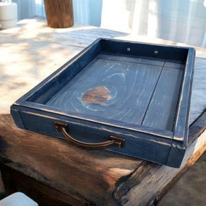 May include: A rectangular wooden tray with a distressed blue finish and a bronze-colored handle. The tray is made of wood and has a rustic, farmhouse style. The tray is approximately 14 inches long and 10 inches wide.